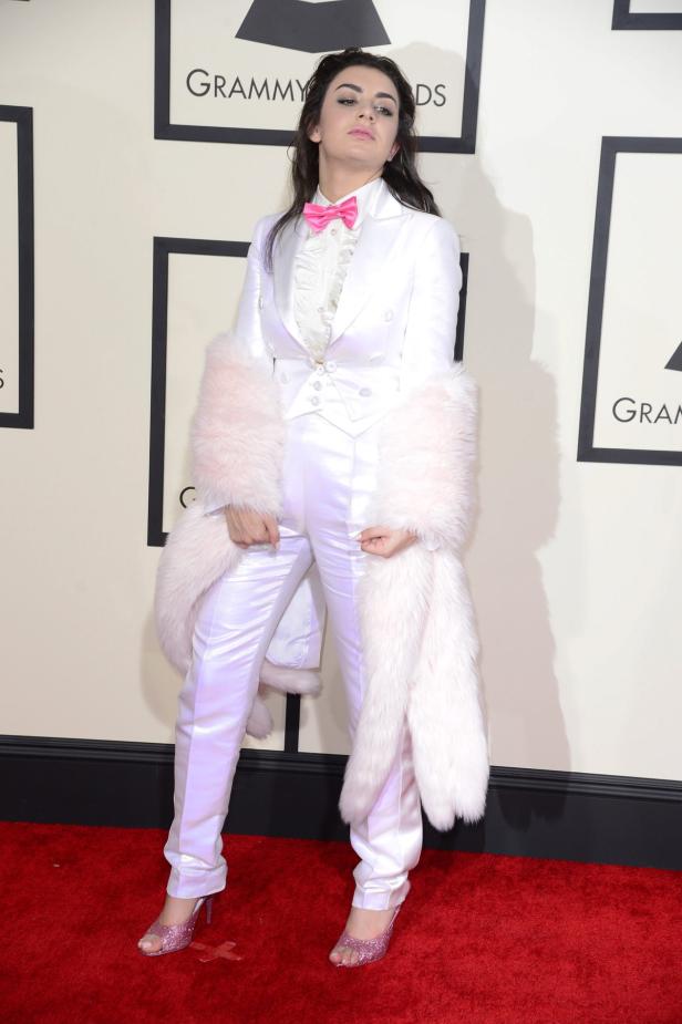 Charli XCX in einem weißen Anzug mit rosa Fliege und Stola auf dem roten Teppich.