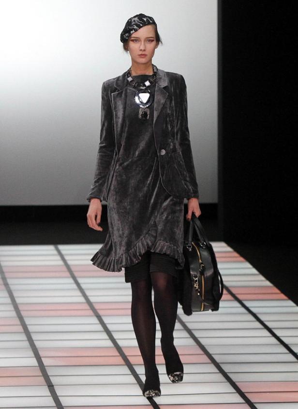 Eine Frau in einem grauen Samtanzug mit Baskenmütze und einer großen Halskette auf dem Laufsteg.