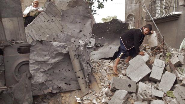 Ein Junge klettert barfuß über Trümmer, während eine ältere Frau zusieht.