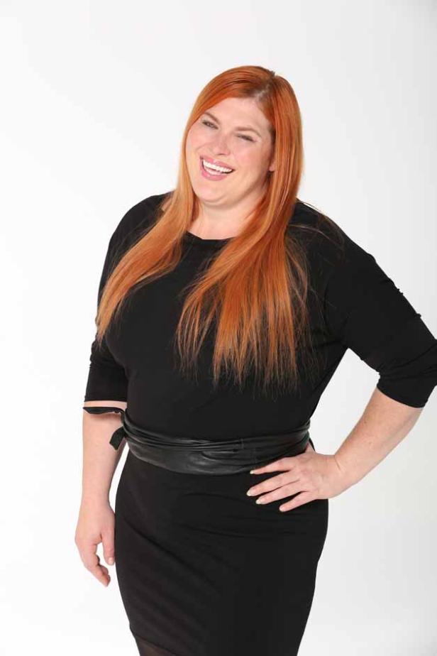 Eine Frau mit roten Haaren lächelt in einem schwarzen Kleid mit Gürtel.