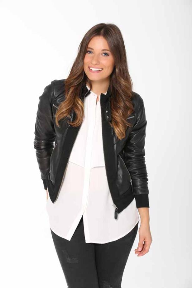 Eine lächelnde Frau mit braunen Haaren trägt eine schwarze Lederjacke und eine weiße Bluse.