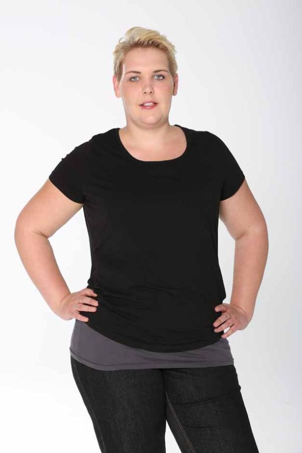Eine Frau mit blonden, kurzen Haaren posiert in einem schwarzen T-Shirt und Jeans vor einem weißen Hintergrund.