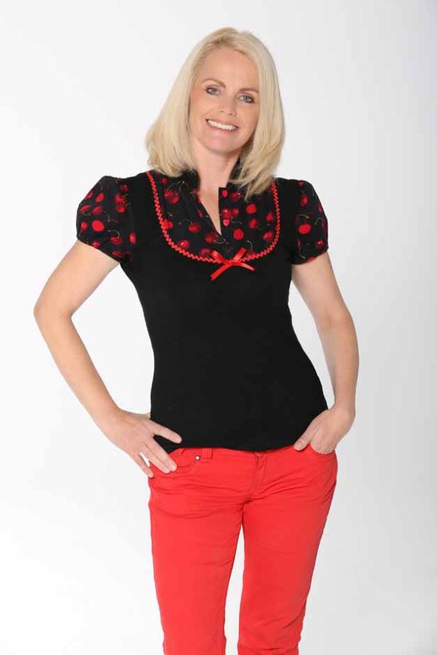 Eine Frau mit blonden Haaren trägt eine schwarze Bluse mit Kirschmuster und rote Jeans.