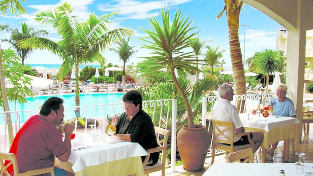 Gäste sitzen auf einer Terrasse mit Blick auf einen Pool und Palmen.