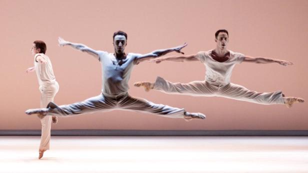 Drei männliche Balletttänzer in weißen Kostümen springen auf einer Bühne.