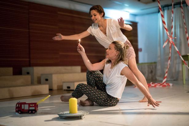 Zwei Frauen interagieren auf einer Bühne mit einer brennenden Kerze und einem Spielzeug-Feuerwehrauto.