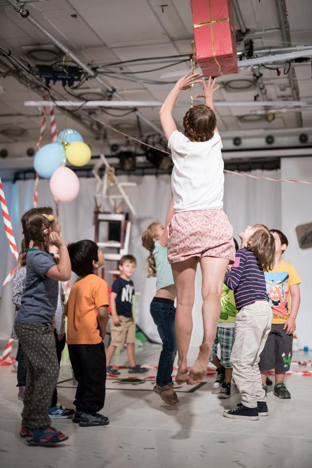 Eine Frau springt, um ein rotes Geschenk von der Decke zu holen, während Kinder zuschauen.