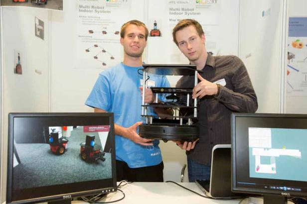 Zwei Männer präsentieren einen mehrstöckigen Roboter auf einem Tisch mit Computermonitoren.