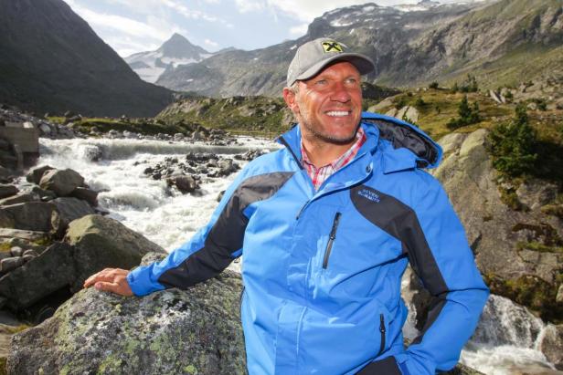 Ein Mann in Berglandschaft mit blau-schwarzer Jacke und Kappe vor einem Fluss.