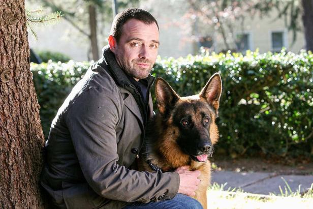 Ein Mann sitzt an einem Baum und umarmt seinen Deutschen Schäferhund.