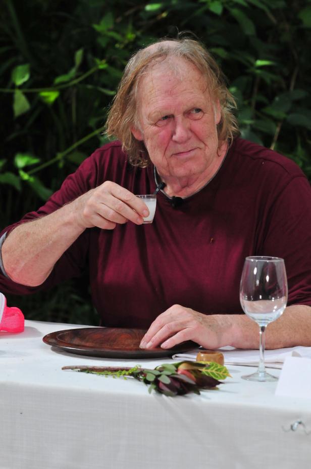Gunter Gabriel Ein Mann mit langen Haaren trinkt aus einem kleinen Glas an einem gedeckten Tisch.