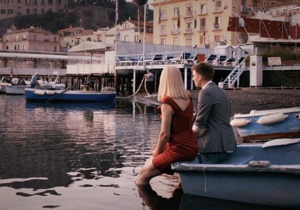 Ein Paar sitzt am Ufer in einem Boot, die Füße im Wasser, mit Blick auf die Küstenstadt.