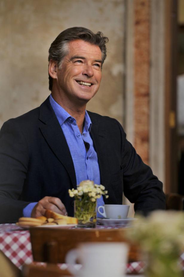 Pierce Brosnan sitzt lächelnd an einem Tisch mit einer Tasse Kaffee und Gebäck.