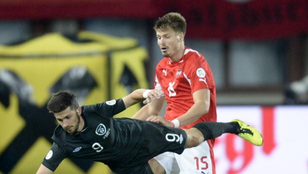 Ein Fußballspieler im schwarzen Trikot wird von einem Spieler im roten Trikot behindert.