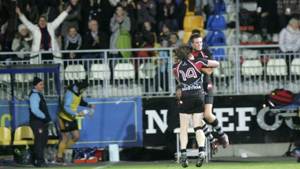 Zwei Rugby-Spieler umarmen sich auf dem Spielfeld, während Zuschauer im Hintergrund jubeln.