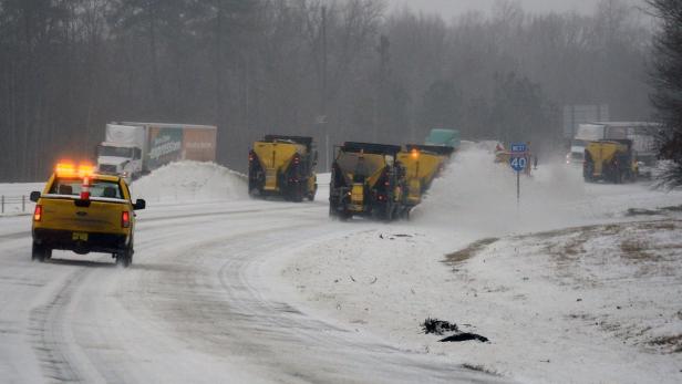 Mehrere gelbe Schneepflüge räumen Schnee auf einer Autobahn, auf der ein Schild „West Route 40“ steht.
