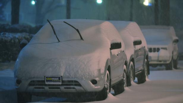 Mehrere unter einer dicken Schneedecke liegende Autos stehen auf einer Straße.