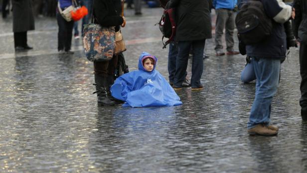 Ein Kind mit blauem Regenponcho sitzt auf einem nassen Platz inmitten von Menschen.