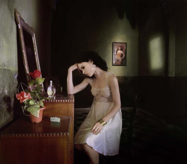 Eine Frau sitzt nachdenklich in einem Zimmer mit einem Spiegel und einer Kommode.