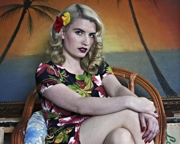 Eine Frau mit blonden Locken und einem Blumenkleid sitzt vor einem Gemälde mit Palmen.