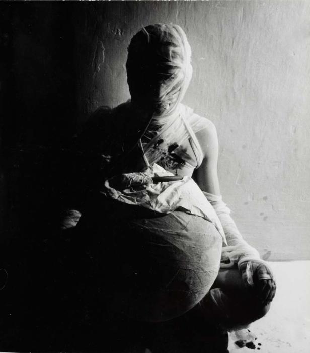 Eine in Bandagen gehüllte Person sitzt mit blutbefleckter Kleidung da.