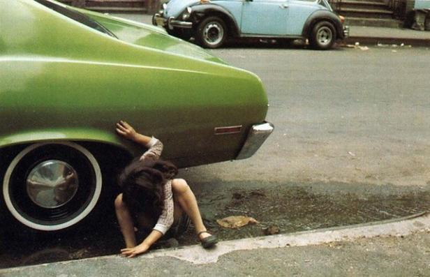 Ein Mädchen kauert unter einem grünen Auto am Straßenrand.