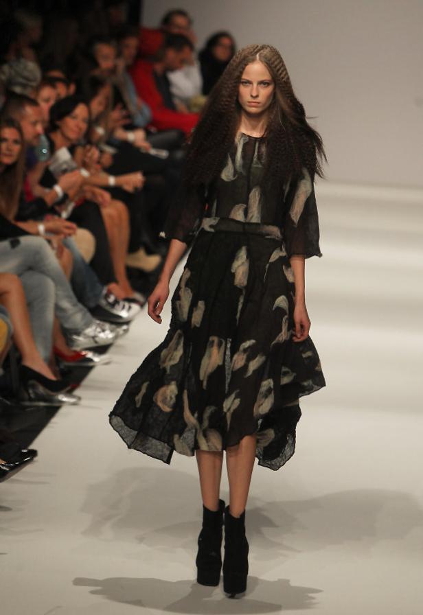 Eine Frau mit gewelltem Haar präsentiert ein gemustertes Kleid und schwarze Stiefeletten auf dem Laufsteg.