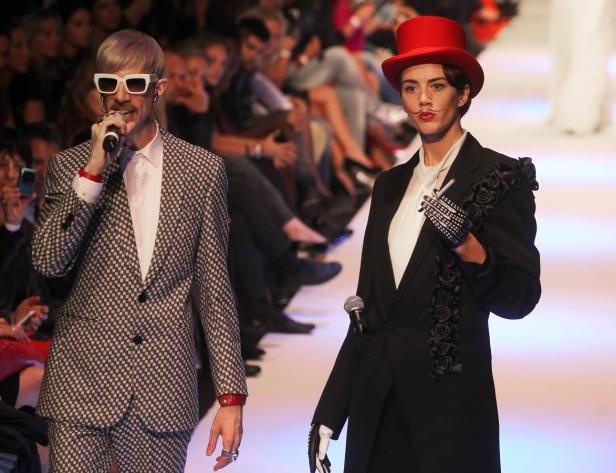 Zwei Personen in extravaganten Anzügen präsentieren Mode auf einem Laufsteg.