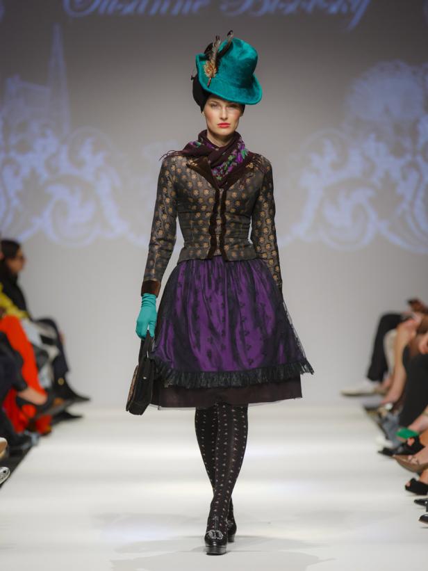 Eine Frau in einem extravaganten Outfit mit Hut läuft über einen Laufsteg.