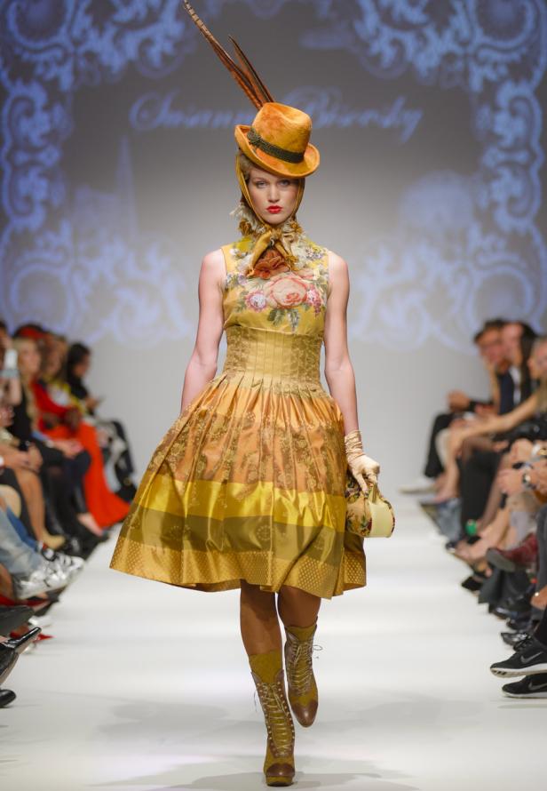 Eine Frau in einem goldenen Kleid mit Blumenmuster und einem Hut mit Federn auf einem Laufsteg.