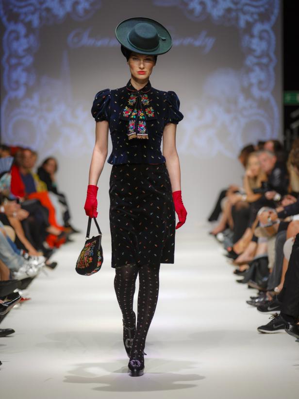 Eine Frau in einem blau-schwarzen Trachtenkleid mit Hut und roten Handschuhen auf einem Laufsteg.