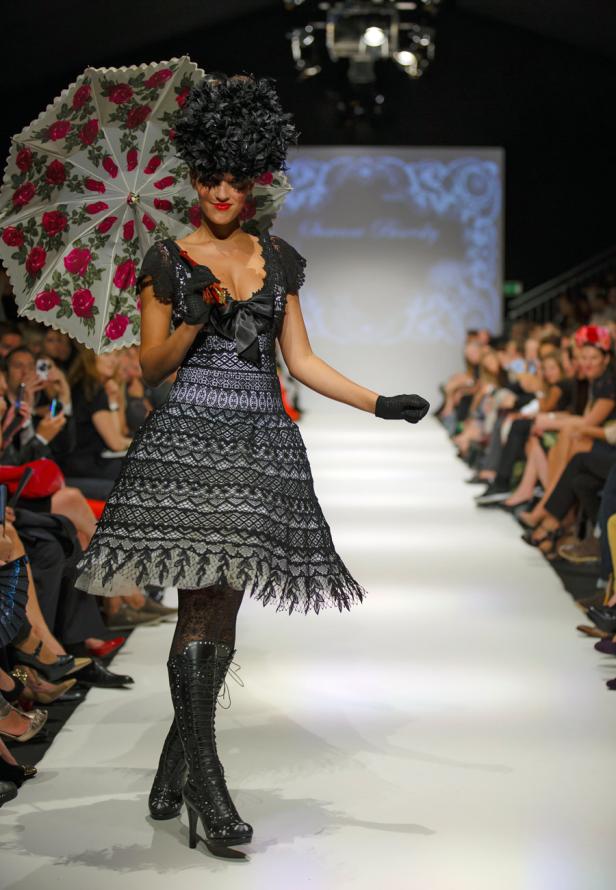 Eine Frau in einem schwarzen Kleid mit Spitze und Hut auf einem Laufsteg mit einem Rosen-Sonnenschirm.