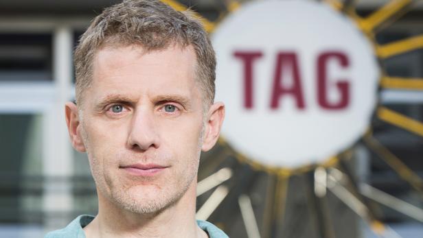 Ein Mann vor einem unscharfen Hintergrund mit dem Wort „TAG“.