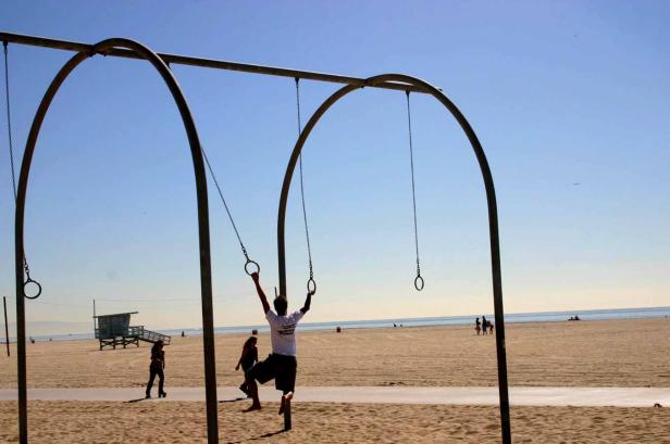 Ein Mann trainiert an Turnringen am Strand, während andere Leute im Hintergrund spazieren gehen.