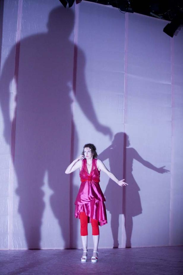 Eine Frau in einem pinkfarbenen Kleid steht auf einer Bühne mit großen Schatten im Hintergrund.