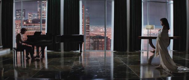 Ein Mann spielt Klavier, während eine Frau in ein Laken gehüllt vor einem Fenster mit Stadtblick steht.