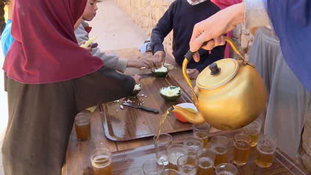Eine Person gießt Tee in Gläser, während Kinder Brot mit Butter essen.