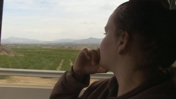 Eine Frau blickt aus dem Fenster eines fahrenden Autos auf eine grüne Landschaft mit Bergen im Hintergrund.