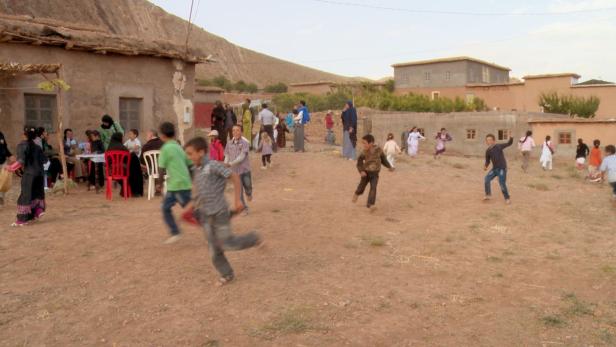 Kinder spielen in einem Dorf, während Erwachsene an Tischen sitzen.