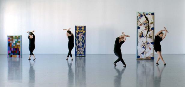 Vier Tänzerinnen in schwarzen Kleidern posieren vor abstrakten Kunstwerken in einem hellen Raum.