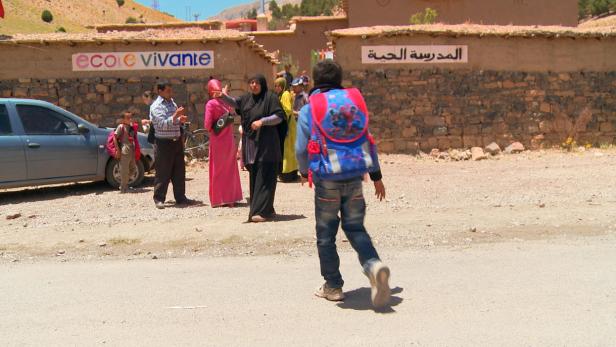 Ein Junge mit einem Rucksack geht auf eine Gruppe von Menschen vor einem Schulgebäude zu.