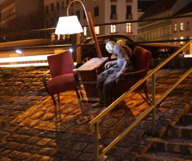 Eine Installation mit Sessel, Teppich, Tisch und einer schlafenden Figur im öffentlichen Raum bei Nacht.
