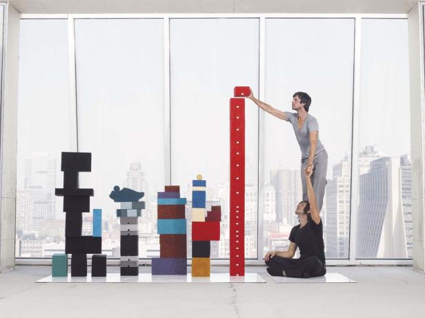 Zwei Männer bauen mit bunten Blöcken eine stilisierte Skyline vor einem Fenster.