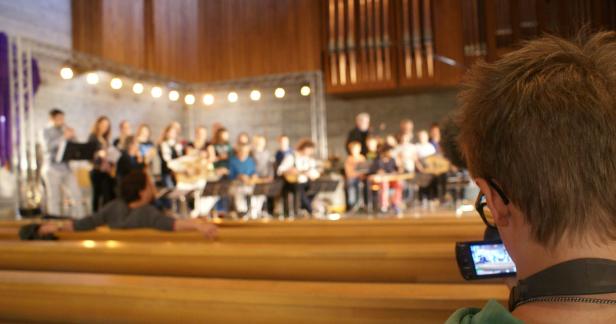 Eine Gruppe von Musikern spielt auf einer Bühne, gefilmt von einer Person im Vordergrund.