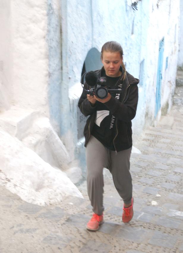 Eine junge Frau filmt mit einer Videokamera in einer Gasse.