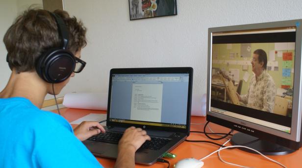 Ein Jugendlicher mit Kopfhörern sitzt vor einem Laptop und einem zweiten Bildschirm, auf dem ein Video läuft.