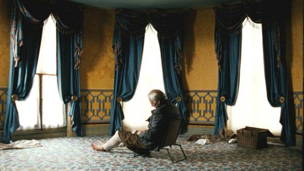 Ein älterer Mann sitzt barfuß in einem Zimmer mit drei Fenstern und liest.