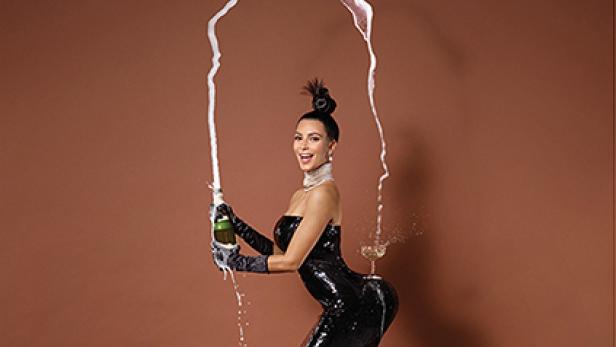 Kim Kardashian öffnet eine Champagnerflasche, wobei ein Glas auf ihrem Hinterteil balanciert.