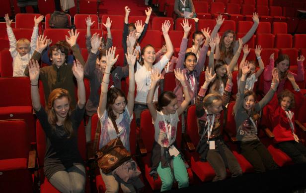Eine Gruppe von Kindern sitzt in einem Theatersaal und hebt die Hände.
