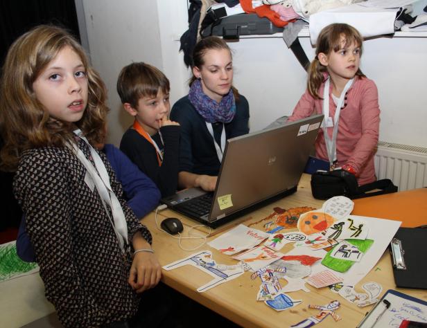 Eine Gruppe von Kindern und eine Frau sitzen an einem Tisch mit einem Laptop und Papierfiguren.